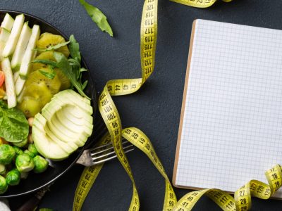 Vegan healthy balanced diet concept. Vegetarian buddha bowl with blank notebook and measuring tape. Сhickpeas, broccoli, pepper, tomato, spinach, arugula and avocado in plate on dark background. Top view